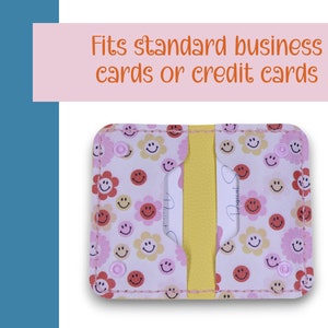 May include: A pink and white card holder with a smiley face and flower pattern. It has a yellow divider and fits standard business cards or credit cards.