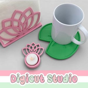 May include: A pink lotus flower-shaped napkin holder, a candle holder, and two green leaf-shaped coasters. A white mug sits on one of the coasters. The items are on a light gray surface. The text "Digicut Studio" is at the bottom.