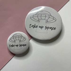 Könnte beinhalten: Zwei weiße runde Knöpfe mit einer schwarzen Umrandung eines UFOs und dem Text "take up space" in schwarzer Schrift.