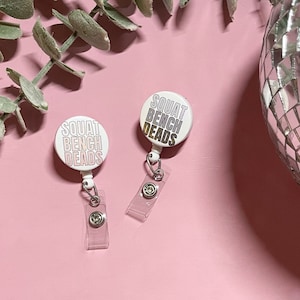 May include: Two white retractable badge holders with a pink background. The badge holders have a white circle with the text "SQUAT BENCH DEADS" in black and pink lettering.