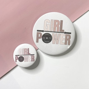 Puede incluir: Dos botones redondos blancos con el texto "GIRL POWER" en letras rosas y marrones. El botón más grande tiene una barra y una placa de peso negras con el texto "BARBELL" y "45 LBS" impreso en él.