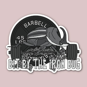 May include: A black and white sticker with a beetle lifting a barbell with chains. The barbell has a weight plate that says "45 LBS" and "STANDARD". The text "Bit by the Iron Bug" is below the barbell.