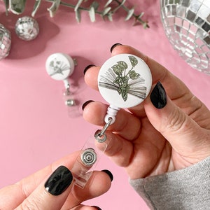 May include: A white retractable badge reel with a botanical illustration of a plant growing from an open book. The badge reel has a clear plastic strap and a metal clip. The background is pink with disco balls and eucalyptus.