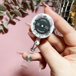 Pode incluir: Um porta-crachá retrátil branco com um design de placa de peso preto e branco. A placa de peso tem o texto "BARBELL STANDARD 45 LBS 20.4 KGS" impresso nela. O design também inclui detalhes florais.