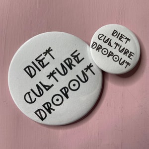 Può includere: Due bottoni rotondi bianchi con testo nero che recita "DIET CULTURE DROPOUT". Il bottone più grande è a sinistra e il bottone più piccolo è a destra.