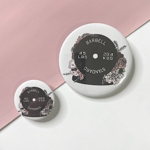 May include: Two round floral weightlifting pins with black centers. The larger pin reads "BARBELL STANDARD 45 LBS 20.4 KGS". The smaller pin reads "BARBELL STANDARD 45 LBS 20.4 KGS".