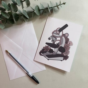 May include: A white card with a pink and gray illustration of a microscope with flowers around it. The microscope has a pink tube and a gray base.