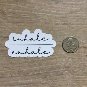 May include: A white sticker with the words "inhale exhale" in black cursive script. The sticker is cloud-shaped and is next to a U.S. quarter coin for size comparison.