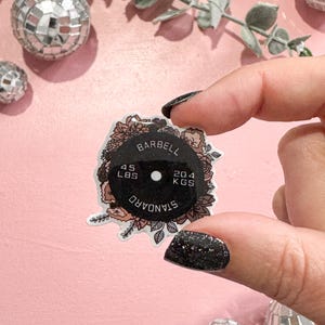 May include: A black and floral barbell weight sticker with the text "BARBELL", "45 LBS", "20.4 KGS", and "STANDARD". The sticker is held against a pink background with disco balls and eucalyptus leaves.