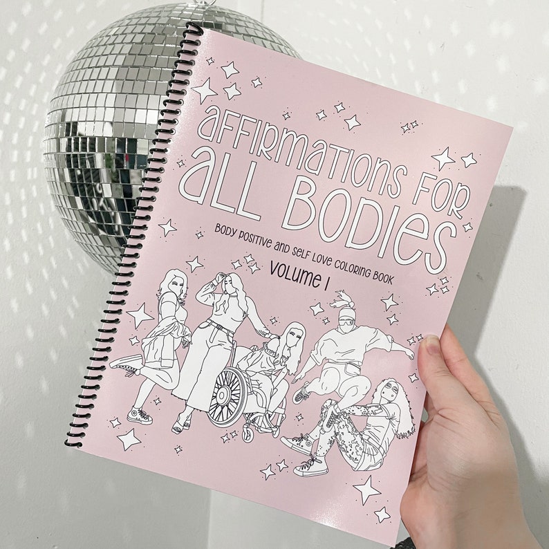 Op de afbeelding: Een roze kleurboek met de titel "Affirmations for All Bodies: Body Positive and Self Love Coloring Book, Volume I". De omslag toont een zwart-witte illustratie van vijf mensen, waaronder een persoon in een rolstoel.