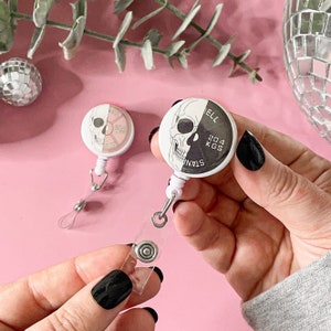 Puede incluir: Un portagajes retráctil con una calavera blanca y negra con el texto "Stay Strong 204 KGS". La calavera es mitad negra y mitad blanca, con el lado negro mostrando una placa de peso.