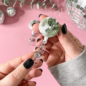 Pode incluir: Um porta-crachá com um design de folha de monstera. O carretel redondo tem um fundo branco com folhas de monstera verdes. Possui uma alça de plástico transparente e um clipe de metal. O fundo é rosa.