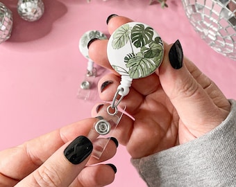 May include: A white and green monstera leaf design badge reel. The retractable reel has a clear plastic strap and a metal clip. The badge reel is round and has a white background with green monstera leaves. The background is pink.