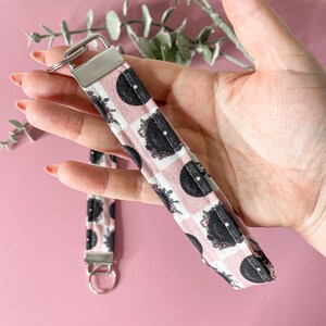 May include: A pink and black fabric key chain with a vinyl record pattern. The key chain has a silver metal ring and a silver metal clasp.