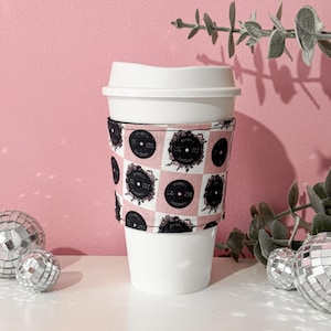 Puede incluir: Una taza de café desechable blanca con tapa blanca y funda de café estampada en rosa y negro. La funda presenta un diseño de tablero de ajedrez con gráficos circulares negros. Bolas de discoteca decorativas y vegetación en el fondo.