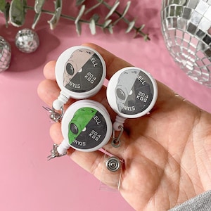 Pode incluir: Três porta-crachás retráteis com desenhos de cabeça de alienígena. Cada porta-crachá tem um fundo preto e branco com o texto "Stan" e "20,4 KGS" impresso nele. As cabeças de alienígenas são de cores diferentes: verde, cinza e bege.