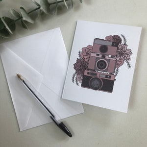 May include: A white card with a stack of three vintage cameras surrounded by pink flowers. The cameras are stacked on top of each other and are all facing the same direction. The flowers are in shades of pink and purple and are arranged around the cameras in a loose, organic way.