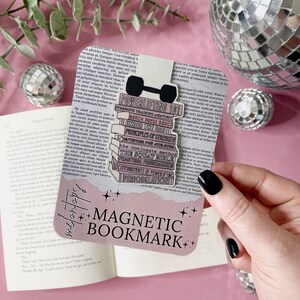Peut inclure: Un marque-page magnétique rose et blanc avec une pile de livres et un haltère. Les livres ont des titres liés à la musculation, tels que "Anatomie de l'entraînement en force", "Bases du développé couché" et "Principes de la force". Le marque-page comporte également le texte "Marque-page magnétique".