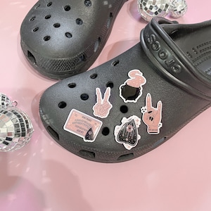 May include: Black and pink witchy themed stickers on a pair of black Crocs. The stickers include a cauldron, a peace sign, a hand with a rock and roll sign, an eye with stars, and a pink Ouija board.