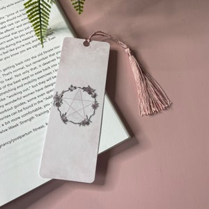 Puede incluir: Un marcapáginas rosa con un borla blanca. El marcapáginas presenta un pentágono con una estrella de cinco puntas en su interior, rodeado de una corona de flores. El marcapáginas está hecho de papel y tiene un acabado mate.