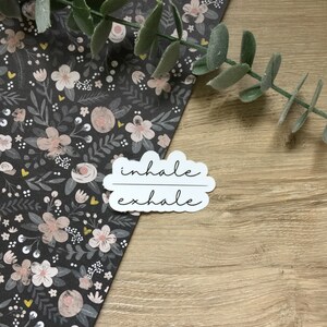 May include: A white sticker with the words "inhale exhale" in a cursive font. The sticker is cloud-shaped and sits on a wooden surface next to a floral patterned paper with pink and white flowers and green leaves.