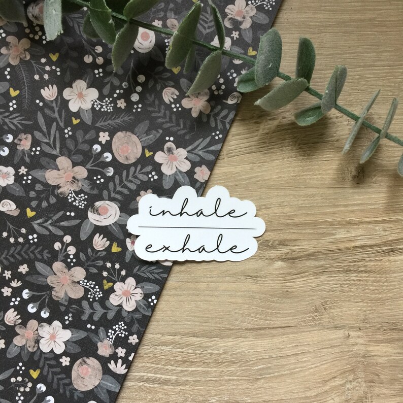 May include: A white sticker with the words "inhale exhale" in a cursive font. The sticker is cloud-shaped and sits on a wooden surface next to a floral patterned paper with pink and white flowers and green leaves.