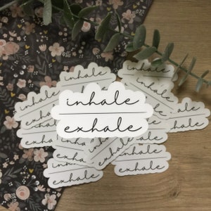 May include: White stickers with a cloud-like shape and the words "inhale" and "exhale" in a handwritten font. The stickers are scattered on a wooden surface with floral patterned paper and greenery in the background.