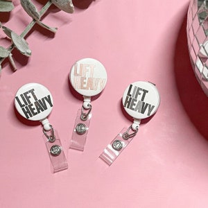 Pode incluir: Três porta-crachás redondos brancos com um gráfico preto e branco "Lift Heavy". Os porta-crachás estão sobre uma superfície rosa.