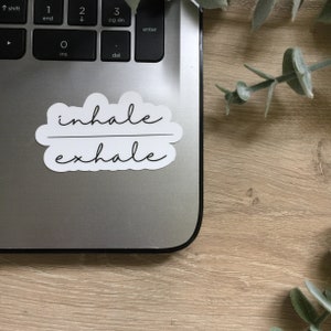 May include: A white sticker with the words "inhale exhale" in a cursive font. The sticker is on a silver laptop, with a wooden surface in the background. Greenery is visible in the top right corner.