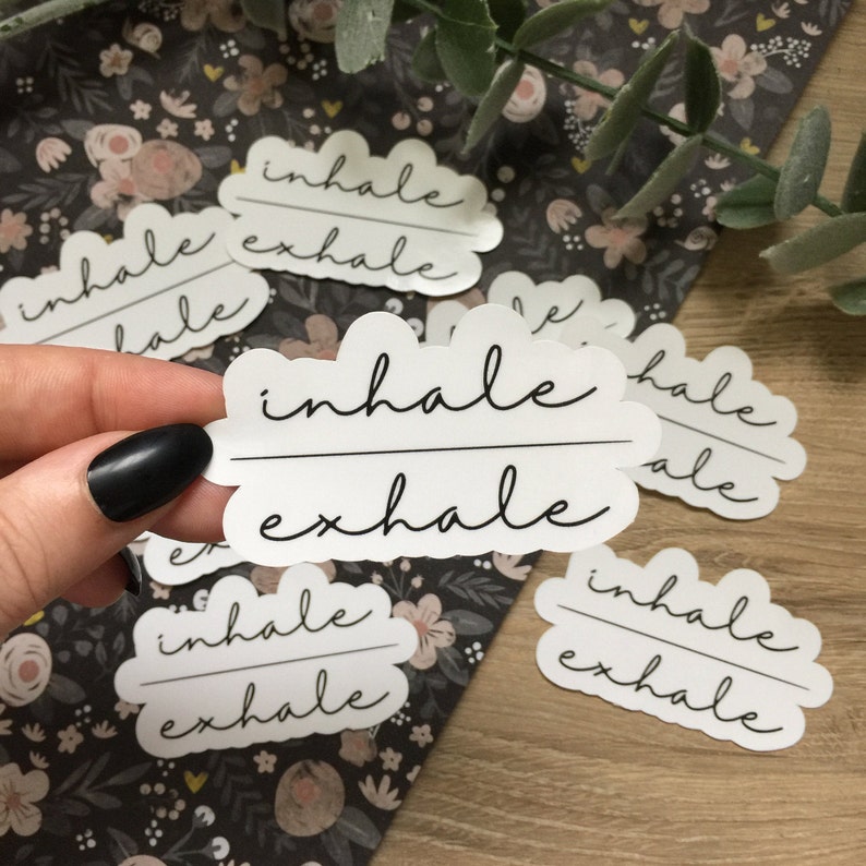 May include: White stickers with the words "inhale exhale" in a handwritten font. The stickers are cloud-shaped and are scattered on a floral patterned background. One hand is holding a sticker.