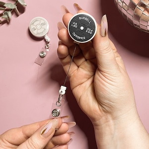 Pode incluir: Dois porta-crachás retráteis de placa de peso preto e branco com um cordão preto. As placas de peso têm o texto "STANDARD BARBELL 45 LBS 20.4 KGS" impresso nelas.