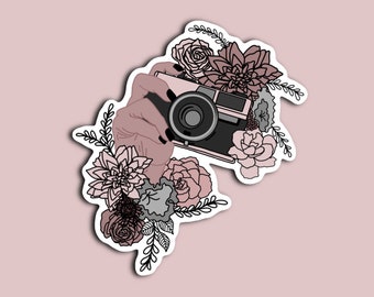 May include: A vintage camera with a black lens is held by a hand surrounded by pink and white flowers. The flowers are drawn in a delicate line art style.