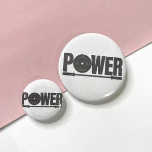 Puede incluir: Dos botones redondos blancos con la palabra "POWER" impresa en negro. El botón más grande presenta una placa de peso negra con el texto "BARBELL" y "45 LBS" y "20.4 KG" impreso en ella. El botón más pequeño tiene una placa de peso negra más pequeña con el texto "BARBELL" impreso en ella.