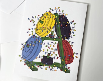 May include: A greeting card featuring a weight rack with colorful weight plates in yellow, blue, black, and red. The rack is decorated with a string of Christmas lights, creating a festive holiday theme.