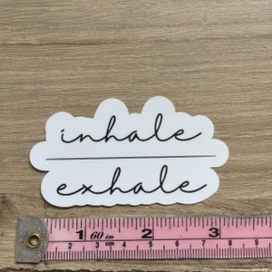 May include: A white sticker with the words "inhale" and "exhale" in black script. The text is divided by a horizontal line. The sticker is on a light brown wooden surface. A pink and white measuring tape is below the sticker.