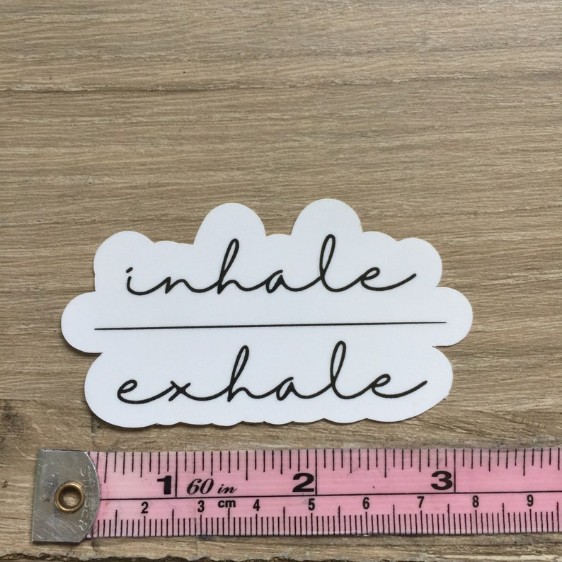 May include: A white sticker with the words "inhale" and "exhale" in black script. The text is divided by a horizontal line. The sticker is on a light brown wooden surface. A pink and white measuring tape is below the sticker.