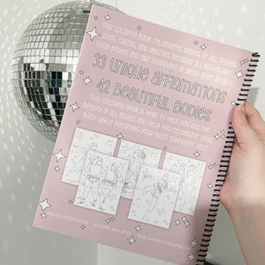 Op de afbeelding: Een roze kleurboek met een spiraalbinding. De omslag heeft een titel die luidt "33 unieke affirmaties 42 prachtige lichamen". Het boek is ontworpen om mensen te helpen hun lichaam te vieren en zelfverzekerder te worden. De omslag bevat ook een paar illustraties van mensen in verschillende poses.