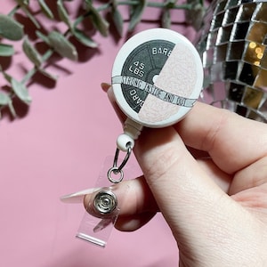 Pode incluir: Um porta-crachá retrátil branco com um design de peso de barra. O peso é preto e cinza, com o texto "45 LBS" e "STRONG INSIDE AND OUT". A outra metade do porta-crachá tem um design de cérebro rosa.