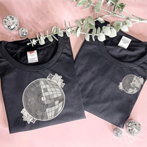 Può includere: Due magliette nere con una grafica bianca di una palla da discoteca e un piatto per pesi. Il piatto per pesi dice "45 LBS" e "BARE" da un lato e "YARD" dall'altro.