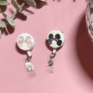 May include: Two white retractable badge holders with a black and pink dumbbell design. The badge holders are on a pink surface.