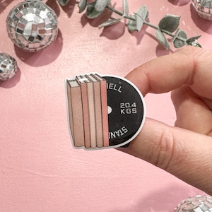 May include: A sticker featuring a stack of books and a weight plate with the text "BELL" and "20.4 KGS". The sticker is held against a pink background with disco balls and eucalyptus leaves.