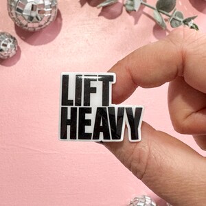 May include: A black and white sticker with the words "LIFT HEAVY" on a pink background. The sticker is held between fingers. Decorative disco balls and greenery are in the background.