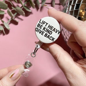 May include: White retractable badge holder with a round white button that says "Lift Heavy Be Kind Give Back" in black text.