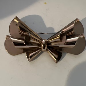 May include: A silver-toned metal brooch in the shape of a bow. The bow is made of multiple overlapping loops and has a smooth, polished finish.