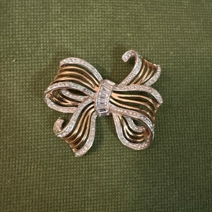 Op de afbeelding: Een vintage strikbroche met een goud- en zilverkleurig ontwerp. De broche heeft een strikvorm met goudkleurig metaal en zilverkleurige metalen accenten. De strik is versierd met heldere, sprankelende kristallen.