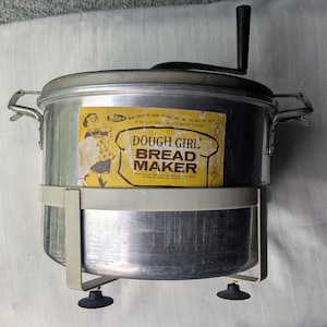 Könnte beinhalten: Vintage Dough Girl Brotbackmaschine aus Aluminium mit schwarzem Griff und gelbem Etikett. Das Etikett trägt die Aufschrift "Dough Girl Bread Maker" mit einer Cartoon-Illustration. Die Brotbackmaschine hat eine silberne Oberfläche und einen weißen Ständer.