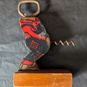 May include: Vintage bottle opener and corkscrew combination. The opener features a wooden base and a decorative figure painted in black and red. The corkscrew is made of metal. A unique barware item.