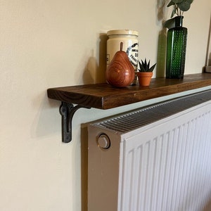 Pu&ograve; includere: Uno scaffale in legno marrone scuro &egrave; montato sopra un radiatore bianco. Lo scaffale &egrave; sostenuto da una staffa in metallo nero. Lo scaffale &egrave; decorato con una pera di legno, una piccola pianta grassa in vaso e un barattolo vintage con un coperchio bianco.
