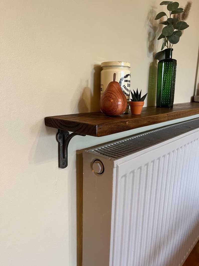 Pu&ograve; includere: Uno scaffale in legno marrone scuro &egrave; montato sopra un radiatore bianco. Lo scaffale &egrave; sostenuto da una staffa in metallo nero. Lo scaffale &egrave; decorato con una pera di legno, una piccola pianta grassa in vaso e un barattolo vintage con un coperchio bianco.