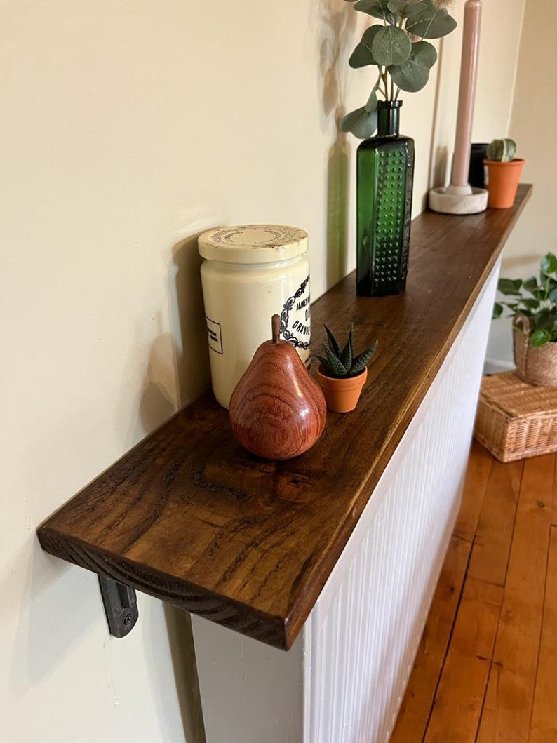 Pu&ograve; includere: Un ripiano in legno marrone scuro con un termosifone bianco dipinto dietro. Il ripiano &egrave; montato alla parete con una staffa in metallo nero. Il ripiano &egrave; decorato con un barattolo di ceramica bianca, una pera di legno, una piccola pianta grassa verde in un vaso di terracotta, una bottiglia di vetro verde con una pianta all'interno e una candela rosa.